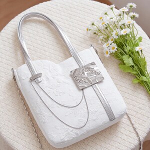 May include: White handbag with silver accents. The bag features a floral pattern, silver chain straps, and a decorative silver clasp. The bag is sitting on a white knitted surface.