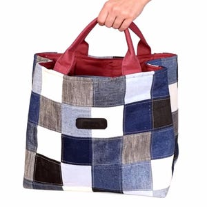 Op de afbeelding: Een patchwork draagtas met een geruit patroon in tinten blauw, wit, grijs en zwart. De tas heeft bordeauxrode handvatten en binnenvoering. Een hand houdt de tas vast. De tas heeft een klein, rechthoekig zwart label.