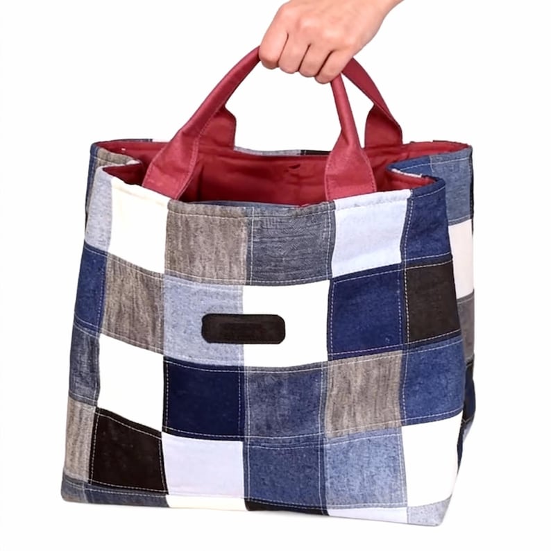 Op de afbeelding: Een patchwork draagtas met een geruit patroon in tinten blauw, wit, grijs en zwart. De tas heeft bordeauxrode handvatten en binnenvoering. Een hand houdt de tas vast. De tas heeft een klein, rechthoekig zwart label.