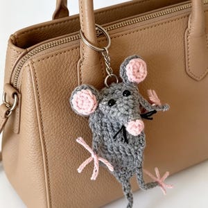 Pode incluir: Um porta-chaves de rato em croché cinzento com orelhas internas, nariz e patas cor-de-rosa, com olhos de missangas pretas e bigodes de fio preto, preso a um anel e corrente de metal prateado numa mala de mão de couro bege.