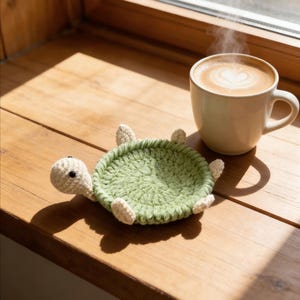 Könnte beinhalten: Ein grüner Häkeltier-Untersetzer in Schildkrötenform mit cremefarbenen Akzenten liegt neben einer weißen Kaffeetasse mit einem herzförmigen Design im Schaum. Der Untersetzer befindet sich auf einer Holzoberfläche in der Nähe eines Fensters.