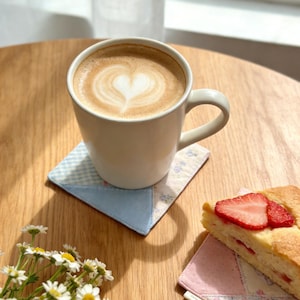 Puede incluir: Una taza blanca con un caf&eacute; con leche, con un dise&ntilde;o en forma de coraz&oacute;n en la espuma, descansa sobre un posavasos de retazos. Una rebanada de pastel de fresa y margaritas est&aacute;n sobre una mesa de madera. El posavasos es azul y blanco.
