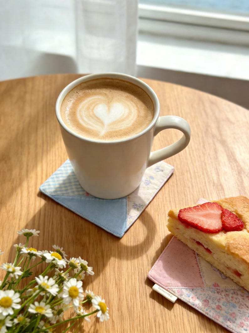 Puede incluir: Una taza blanca con un caf&eacute; con leche, con un dise&ntilde;o en forma de coraz&oacute;n en la espuma, descansa sobre un posavasos de retazos. Una rebanada de pastel de fresa y margaritas est&aacute;n sobre una mesa de madera. El posavasos es azul y blanco.