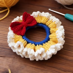 May include: A handmade crochet hair scrunchie with a blue center, yellow and white ruffled trim, and a red bow. The scrunchie is made with yarn and is on a wooden surface. Yarn and a crochet hook are in the background.