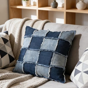 以下が含まれることがあります： 青とライトブルーの色合いのデニムパッチワークの正方形の枕が、グレーのソファの上に置かれています。枕は縁がほつれたデザインです。クリーム色の毛布と他の2つの装飾枕もソファにあります。