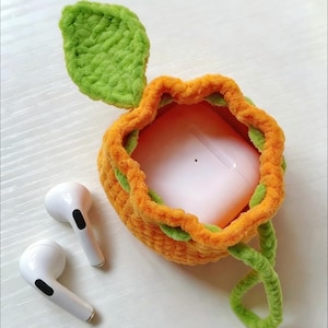 Op de afbeelding: Gehaakte oranje AirPods-hoes met groene blad- en steeld details. De hoes is open en onthult een roze AirPods-hoesje binnenin. Twee witte AirPods liggen naast de hoes op een wit oppervlak. Het ontwerp bootst een vrucht na.