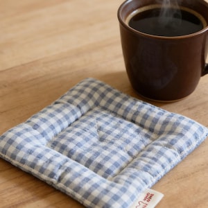 May include: A brown ceramic mug filled with dark coffee, steaming slightly, sits on a blue and white gingham quilted coaster. The coaster has a small tag with text and a heart-shaped graphic.