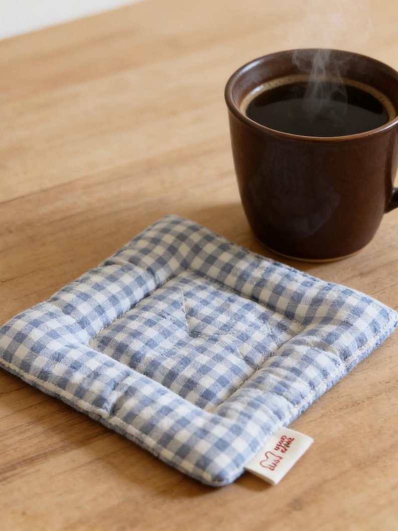 May include: A brown ceramic mug filled with dark coffee, steaming slightly, sits on a blue and white gingham quilted coaster. The coaster has a small tag with text and a heart-shaped graphic.