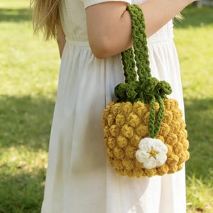 May include: A handmade yellow crochet bag with a green handle and accents, resembling a pineapple. The bag features a white flower embellishment and is held by a person wearing a white dress. The bag's dimensions are approximately 8 inches in height and 6 inches in width.