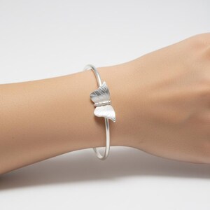 May include: A silver-toned bangle bracelet featuring a central butterfly ornament with etched wing details. The bracelet has a smooth, thin, circular band design that encircles the wrist, showcasing a matte metallic finish throughout the piece.