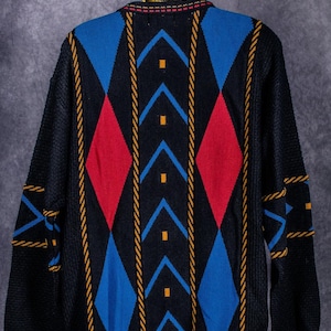 Puede incluir: Suéter negro con un patrón geométrico de rombos rojos y azules, acentuado con líneas amarillas en forma de cuerda. El suéter está colgado en una percha dorada, mostrando un estilo vintage.