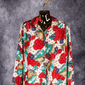 May include: A button-down shirt with a vibrant pattern of red, teal, and tan shapes on a white background. The long-sleeved shirt is displayed on a gold mannequin, hanging from a chain. The design includes floral and nautical elements.