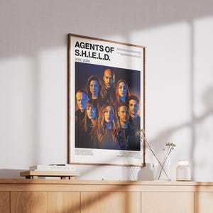 May include: Framed poster featuring the cast of "Agents of S.H.I.E.L.D." with the show's title and the years 2013/2020. The poster is in a wooden frame and is displayed on a wooden cabinet. The background is a wall.