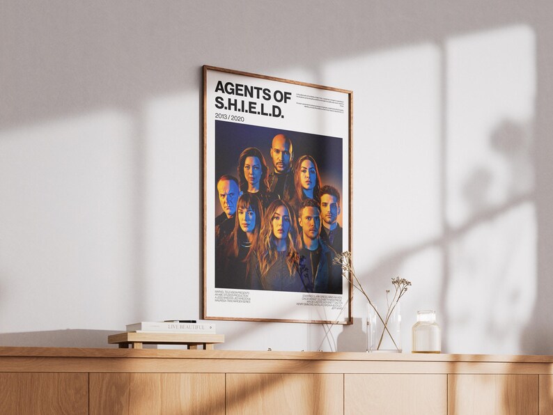 May include: Framed poster featuring the cast of "Agents of S.H.I.E.L.D." with the show's title and the years 2013/2020. The poster is in a wooden frame and is displayed on a wooden cabinet. The background is a wall.