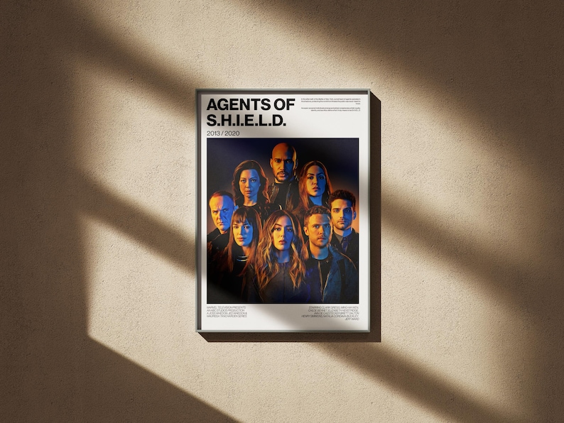 May include: A framed poster featuring the cast of "Agents of S.H.I.E.L.D." The poster has a white border and the title "AGENTS OF S.H.I.E.L.D." is at the top. The poster's background is a beige wall with shadows.