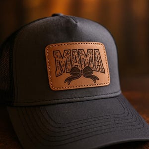 May include: A dark gray trucker hat with a brown leather patch. The patch features the word "MAMA" in a distressed font, with a bow graphic underneath. The hat has a mesh back and a curved brim.