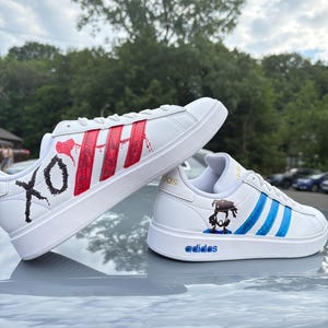 May include: White Adidas sneakers with custom red and blue designs. One shoe features red stripes and the letters "XO" in black. The other shoe has blue stripes and a graphic of a person. The word "adidas" is in blue.