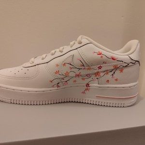 Könnte beinhalten: Weißer Sportschuh mit floralem Design. Der Schuh hat eine weiße Basis mit einem rosa-orangenen floralen Astdesign an der Seite. Die Sohle und die Schnürsenkel sind weiß.