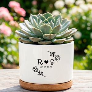 Peut inclure: Un pot de fleurs en céramique blanche pour succulente avec une base en bambou. Le pot présente un motif graphique noir composé de deux fleurs, des lettres R et S séparées par un cœur, et la date 08.10.2026. Une succulente verte est plantée dedans.