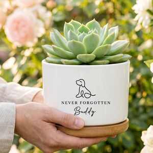 May include: A white ceramic cylindrical planter containing a green succulent. The planter features a black line drawing of a dog and the printed text "NEVER FORGOTTEN" above the name "Buddy" in a script font. It sits on a round bamboo saucer.