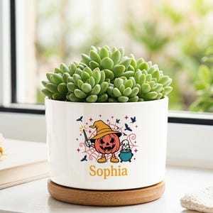 May include: A white cylindrical ceramic planter with a bamboo base, containing a green succulent. The planter features a colourful graphic of a jack-o'-lantern wearing a witch hat, a small ghost, and a cauldron, with the name Sophia printed below.