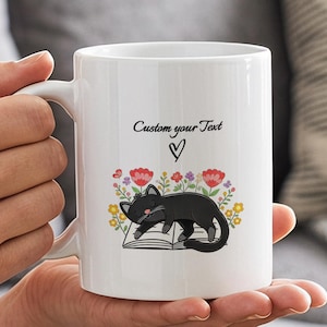 May include: A white ceramic mug featuring a graphic of a black cat sleeping on an open book surrounded by colorful flowers. Above the cat is a small heart icon and the text Custom your Text in a black script font. The mug has a curved handle.