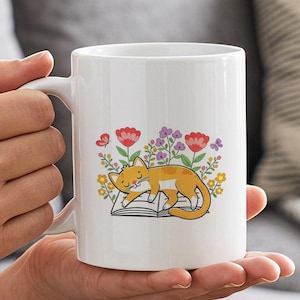 May include: A white ceramic mug featuring a colorful graphic of an orange tabby cat sleeping on an open book, surrounded by a variety of small red, purple, and yellow flowers and two tiny butterflies, all rendered in a simple, clean illustration style.