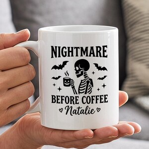 May include: A white ceramic mug featuring a black graphic of a skeleton holding a steaming mug, surrounded by bats and stars. The text reads NIGHTMARE BEFORE COFFEE and the name Natalie, flanked by two small heart symbols in black ink.
