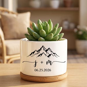 May include: A white ceramic cylindrical planter containing a green succulent. The front features a black graphic of mountain peaks above the text j + m and the date 25.06.2026. The planter rests on a circular light brown wooden base tray.