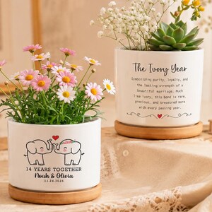 Può includere: Due vasi cilindrici in ceramica bianca su sottovasi in legno. Uno presenta un disegno di due elefanti, il testo 14 YEARS TOGETHER, Noah & Olivia e 11.24.2026. L'altro riporta il testo The Ivory Year e un paragrafo descrittivo.