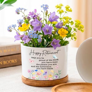May include: A white ceramic cylindrical planter with a bamboo saucer, featuring the text "Happy Retirement" and a floral butterfly design. The planter is filled with purple, yellow, and blue flowers, and an empty matching planter sits beside it.