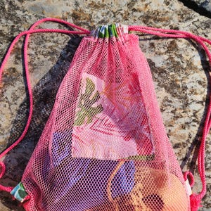Op de afbeelding: Een felroze mesh tas met trekkoord en een paneel van bloemrijke stof aan de voorzijde. De tas heeft bijpassende roze koordhengsels en een geplooide sluiting. Binnenin het doorschijnende mesh zijn een blauw stoffen item en een beige voorwerp zichtbaar.