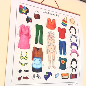 Pode incluir: Um cartaz de boneca de papel com várias roupas e acessórios. As roupas incluem vestidos, tops, calças e chapéus. O cartaz tem o texto "Senhora de Si" e uma assinatura. O cartaz está pendurado numa vara de madeira.