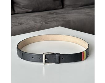 May include: Black leather belt with a silver buckle. The belt features a striped accent in red, yellow, and green. The belt is displayed on a white surface.