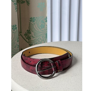 May include: A burgundy belt with a textured snakeskin pattern. The belt has a large, round, silver-colored buckle and a yellow interior. The belt is lying on a white surface.