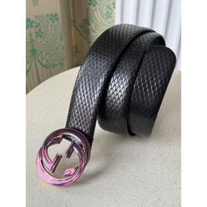 Könnte beinhalten: Schwarzer Ledergürtel mit einem strukturierten Rautenmuster. Der Gürtel hat eine große, schillernde Schnalle mit einem Doppel-G-Logo. Der Gürtel ist auf einer hellen Oberfläche aufgerollt.