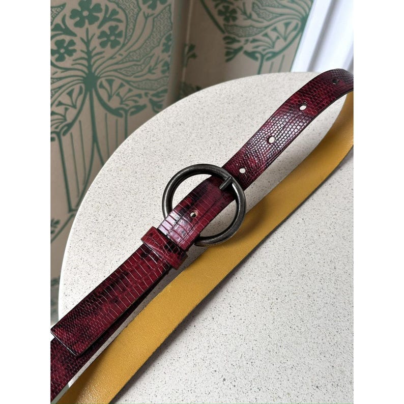 May include: A burgundy faux-snakeskin belt with a large, dark metal ring buckle. A second, mustard-yellow belt is partially visible beneath the burgundy belt. The belts are displayed on a light-colored, speckled surface.