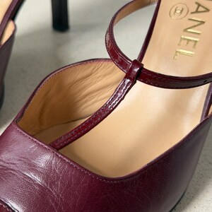 May include: A pair of burgundy leather T-strap heels featuring a glossy patent leather strap across the instep. The interior sole is beige and displays the gold Chanel logo. The shoes have a closed toe design and a slim heel structure.