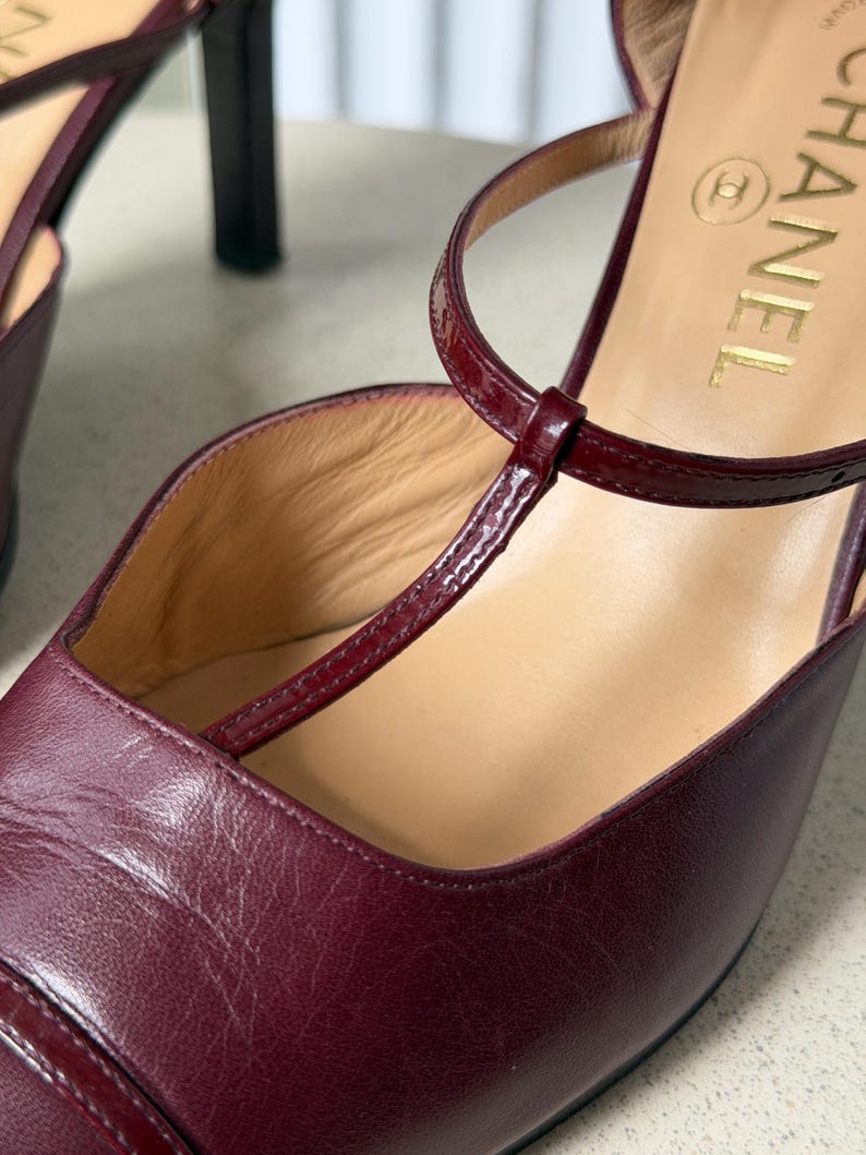 May include: A pair of burgundy leather T-strap heels featuring a glossy patent leather strap across the instep. The interior sole is beige and displays the gold Chanel logo. The shoes have a closed toe design and a slim heel structure.