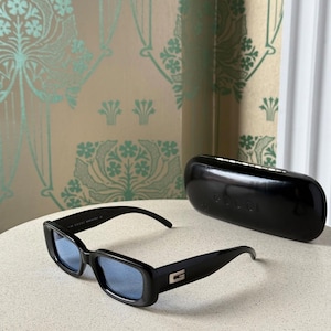 May include: Black rectangular sunglasses with blue lenses and a matching black case. The sunglasses have a silver accent on the temples and are displayed on a light-colored surface.