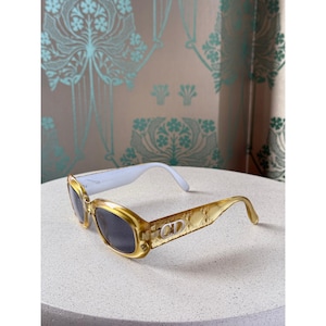May include: Gold-toned sunglasses with dark lenses. The frames have a textured pattern and the brand's initials, "CD", are visible on the temples. The arms are a light blue color. The sunglasses are displayed on a light-colored surface.