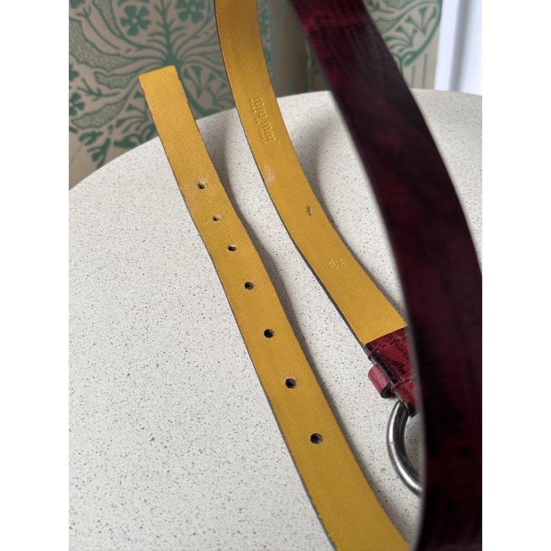 May include: Close-up of a burgundy leather belt with a silver-tone ring buckle. The belt has a yellow interior and several holes for adjustment. The belt is on a white surface.