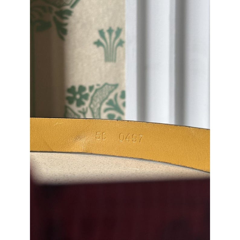 May include: Close-up of a yellow leather belt. The belt's underside displays the stamped numbers "5C 0497". The background includes a patterned fabric and a white curtain, providing a neutral backdrop for the belt's details.