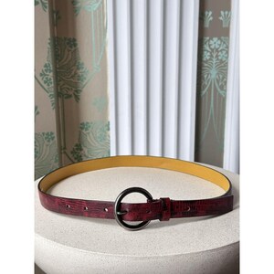 May include: A burgundy belt with a textured finish and a circular, dark metal buckle. The belt has a yellow interior and is displayed on a white, rounded surface. The background features patterned curtains and a white, slatted window covering.