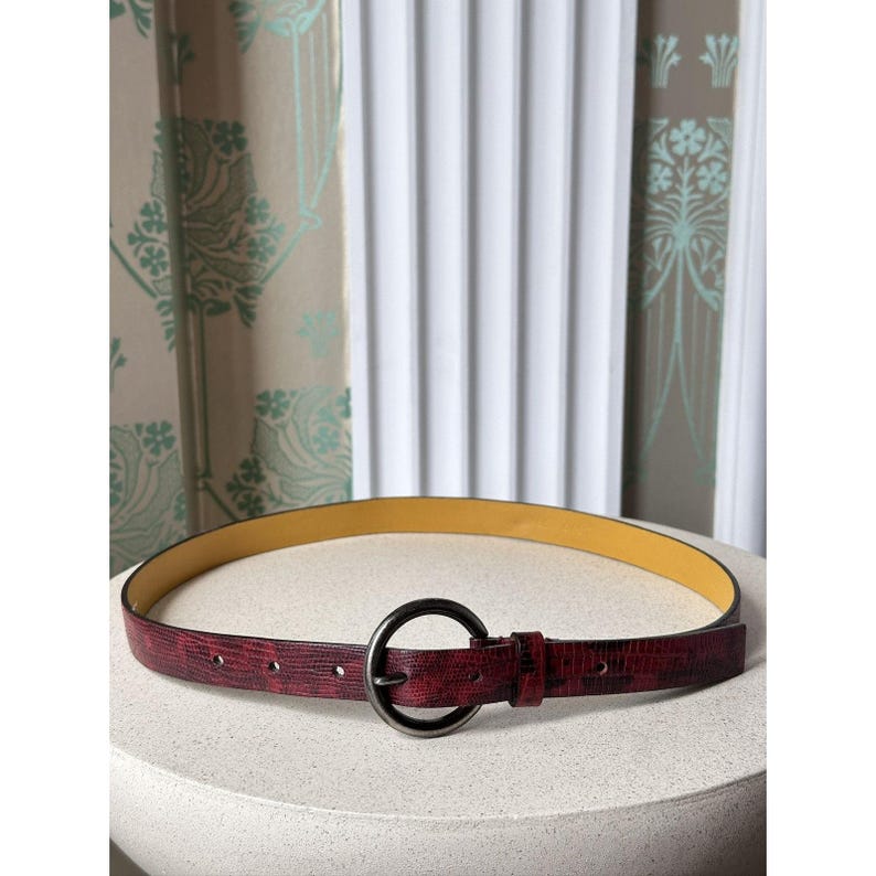 May include: A burgundy belt with a textured finish and a circular, dark metal buckle. The belt has a yellow interior and is displayed on a white, rounded surface. The background features patterned curtains and a white, slatted window covering.