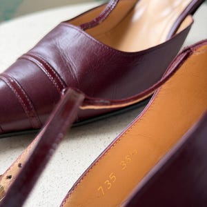 May include: A pair of dark burgundy leather T-strap heels with a pointed toe and thin ankle straps. The interior lining is tan leather, featuring the stamped text 735 38 1/2. The shoes have a glossy finish and visible stitching along the toe cap.