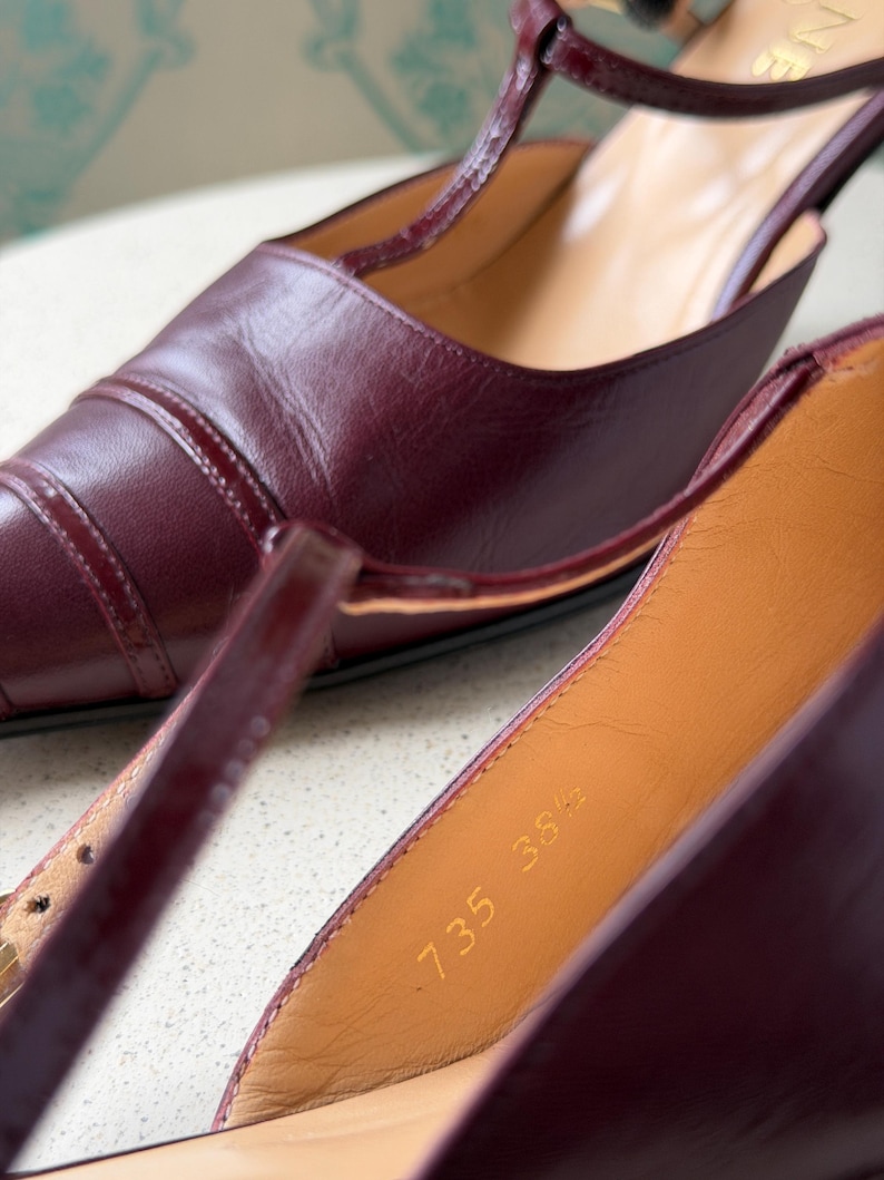 May include: A pair of dark burgundy leather T-strap heels with a pointed toe and thin ankle straps. The interior lining is tan leather, featuring the stamped text 735 38 1/2. The shoes have a glossy finish and visible stitching along the toe cap.