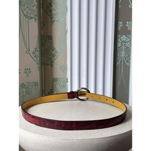 May include: A red textured belt with a silver-colored circular buckle. The belt has a yellow interior and is resting on a white surface. The background includes a patterned curtain and a white wall.