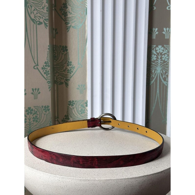 May include: A red textured belt with a silver-colored circular buckle. The belt has a yellow interior and is resting on a white surface. The background includes a patterned curtain and a white wall.