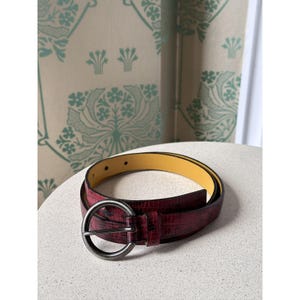 May include: A burgundy belt with a textured, reptile-like pattern. The belt has a large, round, silver-colored buckle and a yellow interior. The belt is displayed on a white surface.